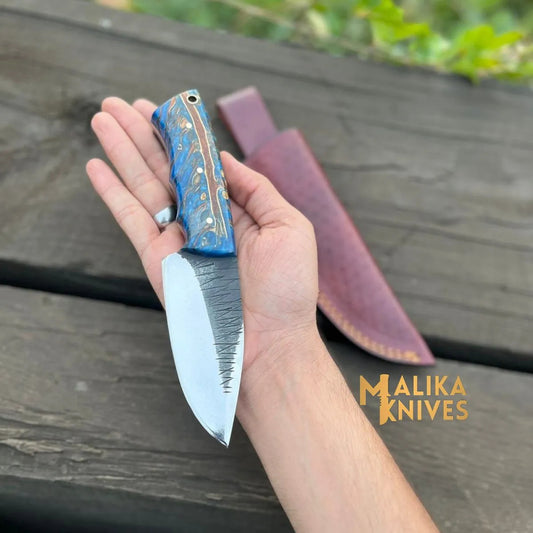 Hand holding Malika Knives custom D2 steel skinner knife with blue resin copper swirl handle blade-up, burgundy leather sheath