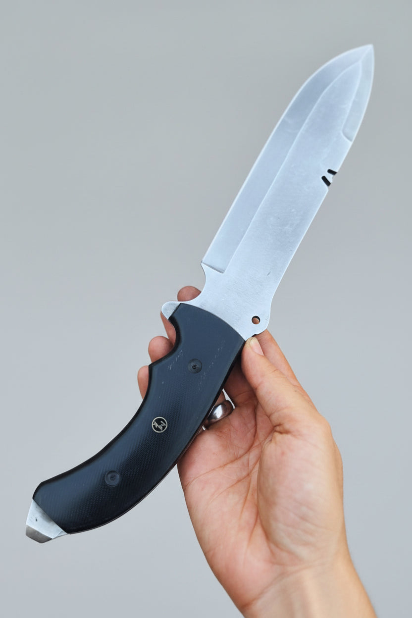 A hand holding a custom spear-point Bowie knife with a black G10 Micarta handle and a silver D2 steel blade, alongside a brown Cowhide leather sheath.