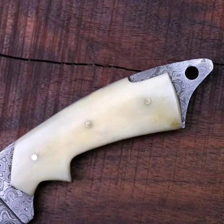 Close-up of camel bone handle on Damascus skinner knife showing ergonomic finger groove, steel pins, and Damascus bolster detail on walnut wood surface
