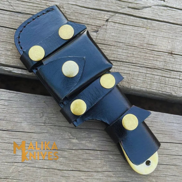 Black leather belt sheath with brass studs for camel bone skinner knife laid flat on weathered wood, showing Malika Knives branding and camel bone handle tip