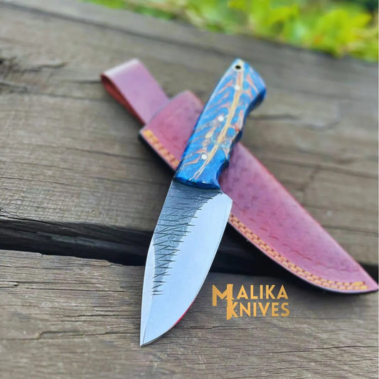 Malika Knives custom D2 steel skinner knife with blue resin copper swirl handle and file-worked spine laid on weathered wood beside hand-tooled burgundy leather sheath

