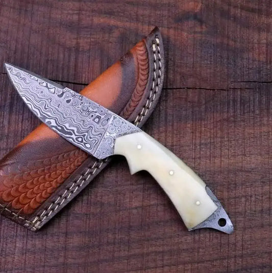Handmade Damascus skinner knife with camel bone handle resting against tooled cowhide leather sheath on dark wood, showing full knife and sheath profile