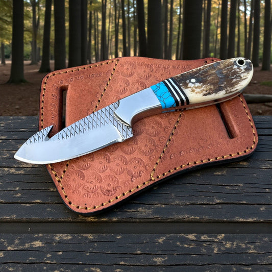 D2 Steel Cowboy Gut Hook Knife with 5mm thick blade and stag handle on leather pancake sheath in forest setting.
