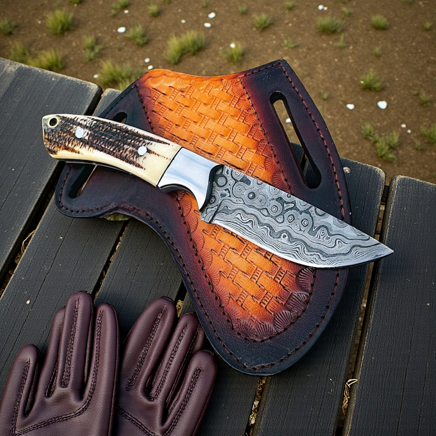 Damascus cowboy knife with antler handle resting on a basket-tooled pancake leather sheath beside brown gloves on an outdoor bench.