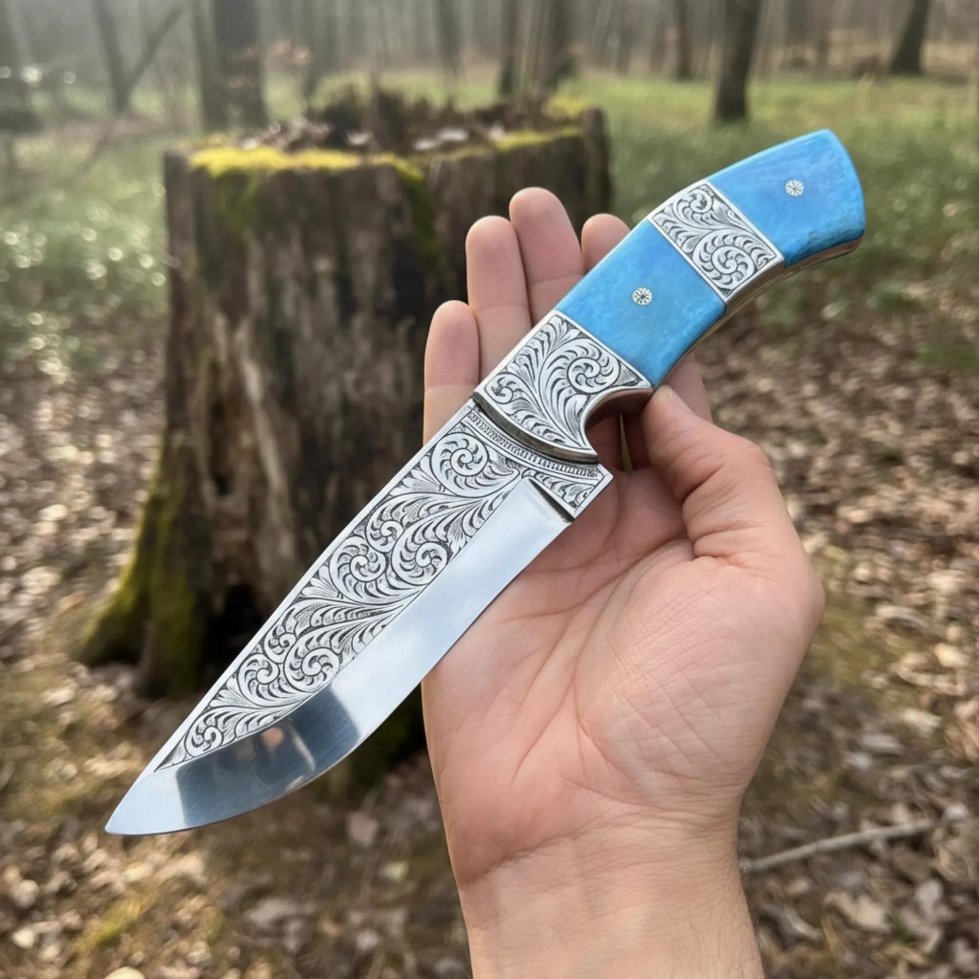 Hand-engraved hunting knife with a Tinted Camel bone handle, held in hand against a wooden background.