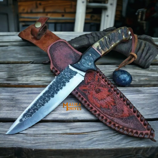 hand forged d2 hunting knife with bull horn handle and engraved leather sheath on wooden outdoor surface