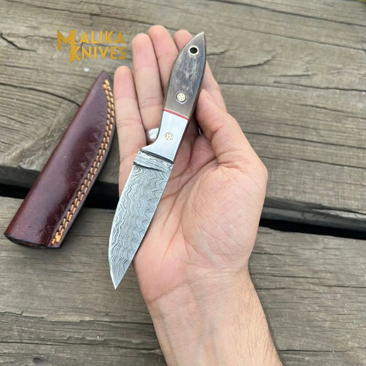 Malika Knives Damascus steel skinner knife with ram horn handle held in hand, showing wavy Damascus pattern blade and leather sheath